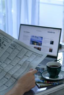 a person reading a newspaper next to a laptop by Anna Keibalo courtesy of Unsplash.