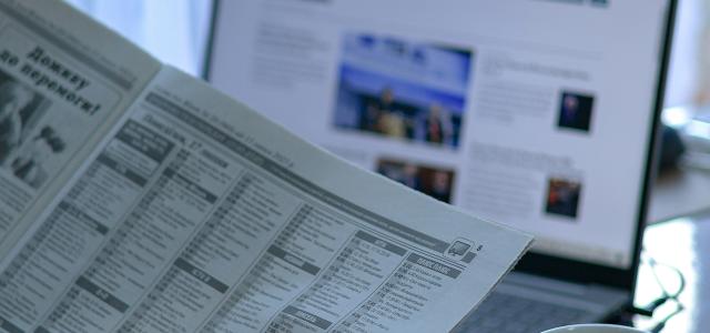 a person reading a newspaper next to a laptop by Anna Keibalo courtesy of Unsplash.